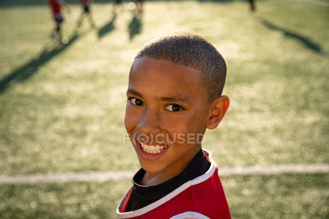 Retrato de cerca de un joven futbolista mestizo usando una tira de equipo, parado en un campo de juego bajo el sol, girando hacia la cámara y sonriendo, con compañeros de equipo en el fondo - foto de stock