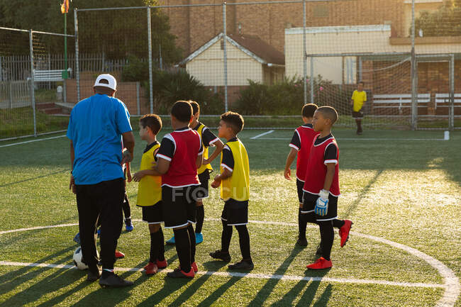 Rear view of a mixed race male soccer coach and instructing a multi-ethnic group of boy soccer players on a soccer pitch in the sun during a soccer training session — Stock Photo