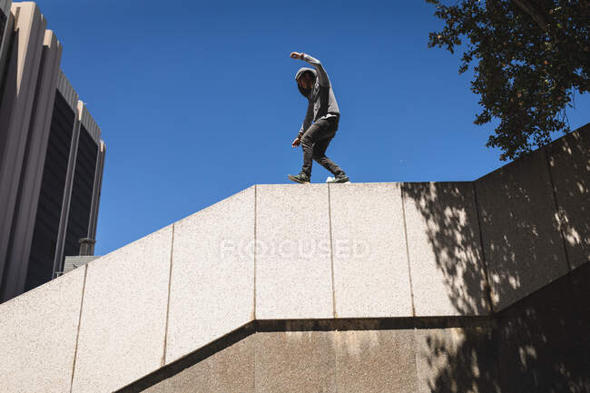 Vista lateral de un hombre caucásico practicando parkour junto al edificio en una ciudad en un día soleado, caminando sobre una barandilla de escaleras de hormigón. - foto de stock