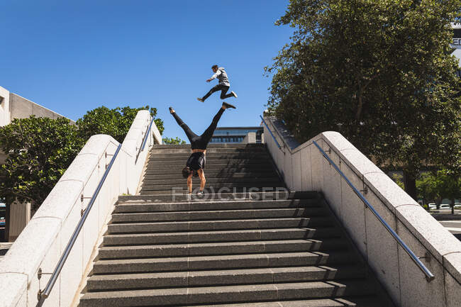 Vista posteriore di un uomo caucasico che pratica il parkour vicino all'edificio in una città in una giornata di sole, facendo stand mentre un altro uomo salta sullo sfondo. — Foto stock