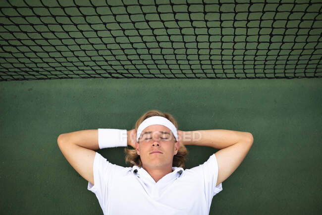 Un hombre caucásico vistiendo blancos de tenis pasando tiempo en una cancha jugando al tenis en un día soleado, tendido en un suelo con los ojos cerrados junto a una red - foto de stock