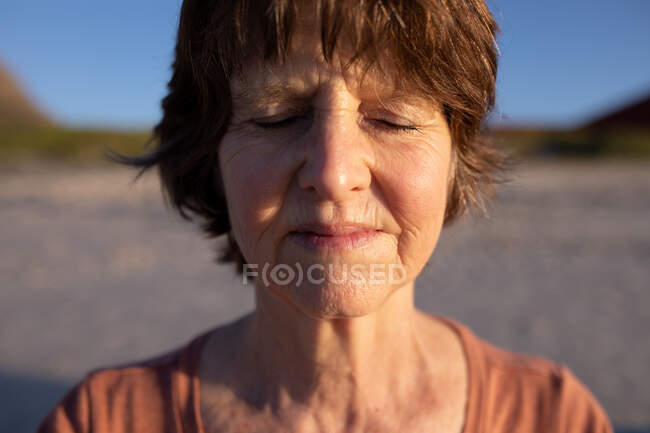 Vista frontal close-up da mulher caucasiana sênior desfrutando de tempo livre em uma praia em um dia ensolarado, meditando com os olhos fechados. — Fotografia de Stock