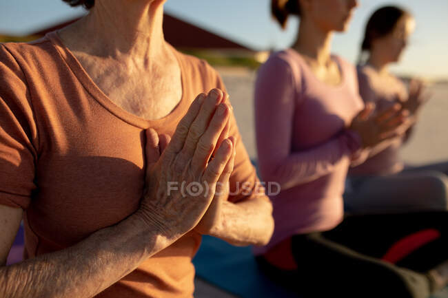 Vue latérale au milieu d'un groupe d'amies profitant de la détente sur une plage par une journée ensoleillée, pratiquant le yoga assis et méditant, les mains dans la prière. — Photo de stock