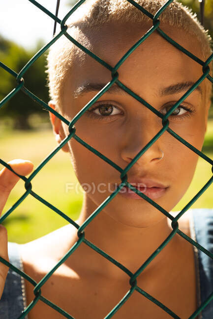 Retrato de mujer alternativa de raza mixta con pelo corto y rubio en la ciudad en un día soleado, mirando a través de una cerca de eslabones de cadena. Mujer urbana independiente sobre la marcha. - foto de stock