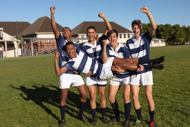 Vista frontal de un grupo de jugadores de rugby masculinos multiétnicos adolescentes que usan tira de equipo azul y blanco, celebrando una victoria, llevando a uno de sus jugadores y animando con los brazos en el aire, de pie en un campo de juego durante un partido - foto de stock