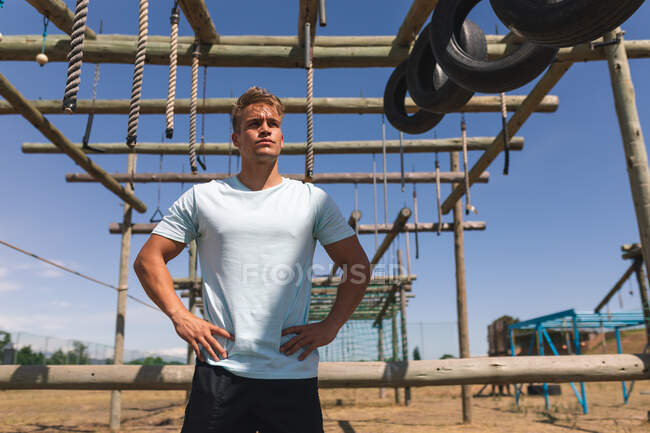 Kaukasische männliche Fitnesstrainerin bei einem Bootcamp an einem sonnigen Tag, die Hände auf den Hüften unter dem Klettergerüst einer Dschungel-Turnhalle mit Hängeseilen stehend — Stockfoto