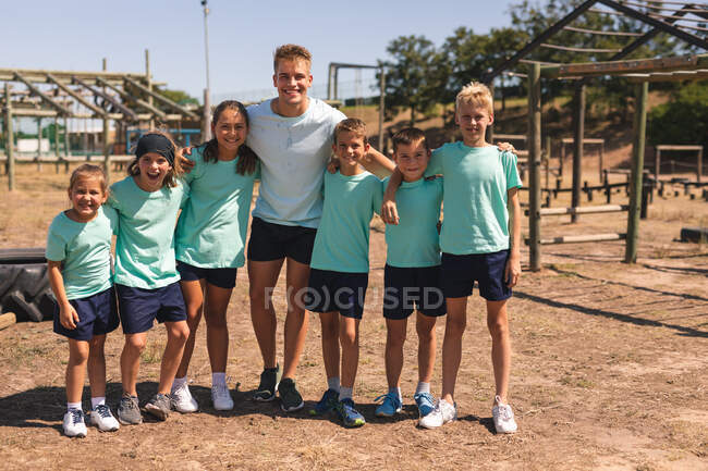 Portrait of a happy group of Caucasian boys and girls and a Caucasian male fitness coach having fun together at a boot camp on a sunny day, with arms around each other, smiling to camera — Stock Photo