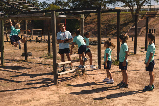Un allenatore di fitness maschile caucasico istruisce un gruppo di ragazzi e ragazze caucasici in un campo di scarponi in una giornata di sole tutti indossando magliette verdi e pantaloncini neri, arrampicandosi su una palestra della giungla e usando i bar delle scimmie. — Foto stock