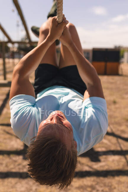 Entrenador de fitness masculino caucásico en un campamento de entrenamiento en un día soleado, colgando boca abajo sosteniendo una cuerda con sus manos y pies en un gimnasio de la selva - foto de stock