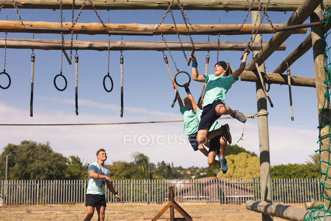 Ein kaukasischer männlicher Fitnesstrainer, der an einem sonnigen Tag zwei kaukasische Jungen in einem Bootcamp unterrichtet, grüne T-Shirts und schwarze Shorts trägt, auf einer Dschungelturnhalle klettert und an Ringen hängt — Stockfoto
