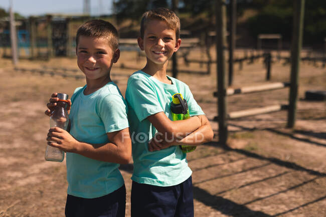 Retrato de dois garotos caucasianos em um acampamento de botas em um dia ensolarado, de pé ao lado uns dos outros segurando garrafas de água vestindo camisetas verdes e shorts pretos olhando para a câmera e sorrindo — Fotografia de Stock