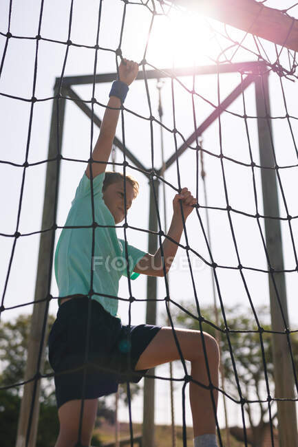 Caucasian boy at a boot camp on a sunny day, climbing up a net on a climbing frame, wearing a blue sweatband, green t shirt and black shorts, backlit — Stock Photo