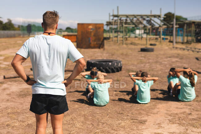 Задний вид на кавказского тренера мужского пола, стоящего и смотрящего на группу кавказских мальчиков и девочек в лагере в солнечный день, делающего хрусты или сидящего, все в зеленых футболках и черных шортах — стоковое фото