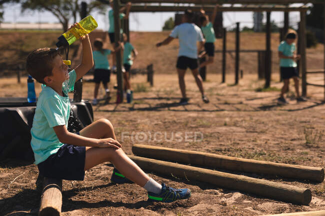 Niño caucásico en un campamento de entrenamiento en un día soleado, sentado en un tronco y vertiendo agua de una botella de agua sobre su cabeza para refrescarse, con un entrenador masculino y otros niños haciendo ejercicio en el fondo - foto de stock