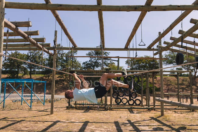 Кавказский тренер по фитнесу в тренировочном лагере в солнечный день, висит вверх ногами, держа веревку руками и ногами на тренажерном зале в джунглях — стоковое фото