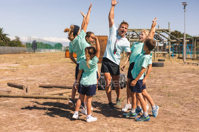 Um grupo feliz de meninos e meninas caucasianos e um treinador de fitness masculino caucasiano se divertindo juntos em um campo de treinamento em um dia ensolarado, gritando por motivação e sorrindo com os braços no ar — Fotografia de Stock