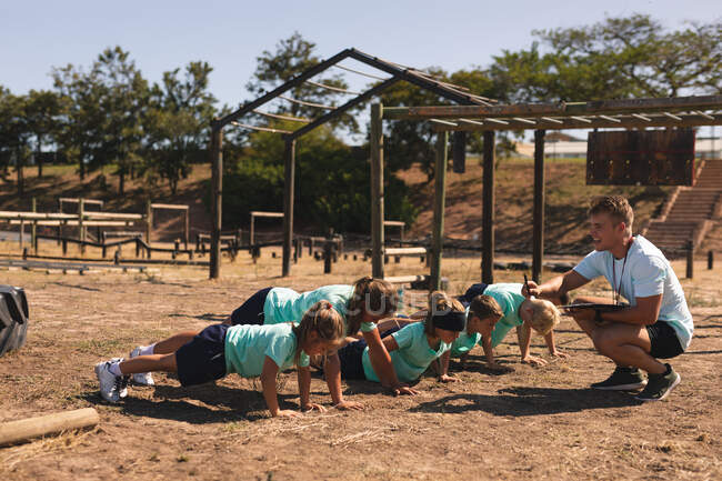 A Caucasian male fitness coach squatting and instructing a group of Caucasian boys and girls at a boot camp on a sunny day doing push ups, all wearing green t shirts and black shorts — Stock Photo