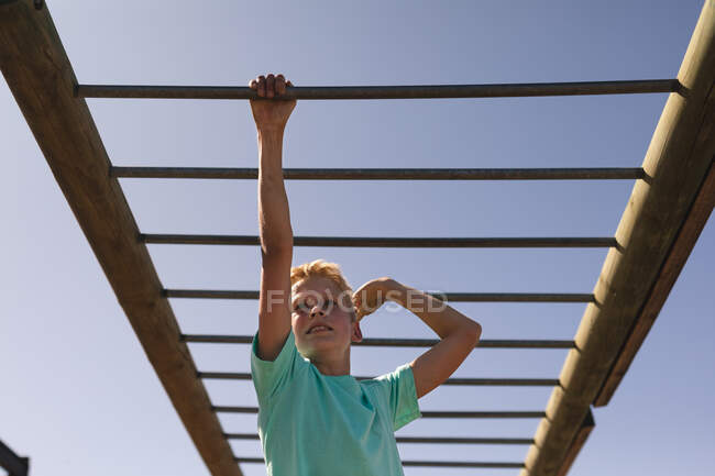 Ragazzo caucasico sorridente con i capelli biondi in un campo di addestramento in una giornata di sole, indossando maglietta verde, su una palestra della giungla appesa alle sbarre delle scimmie contro un cielo blu — Foto stock