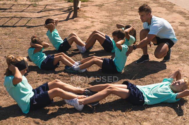 Un entraîneur de conditionnement physique masculin caucasien s'agenouillant et instruisant un groupe de garçons et de filles caucasiens à un camp d'entraînement par une journée ensoleillée, faisant des craquements ou s'asseyant, tous portant des t-shirts verts et des shorts noirs — Photo de stock