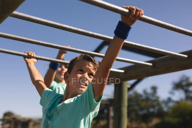 Garçon caucasien souriant aux cheveux bruns dans un camp d'entraînement par une journée ensoleillée, portant un t-shirt vert, sur une salle de gym de jungle suspendue aux barres de singe contre un ciel bleu, un autre garçon derrière lui en arrière-plan — Photo de stock