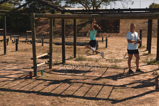 A Caucasian male fitness coach instructing a Caucasian girl at a boot camp on a sunny day wearing green t shirt and black shorts, on a jungle gym hanging from the monkey bars — Stock Photo