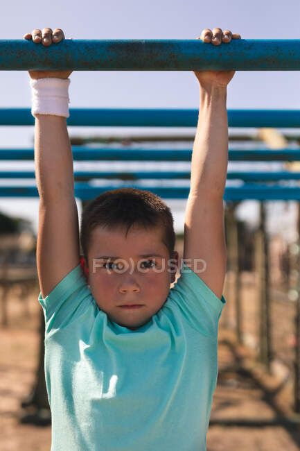 Retrato de menino caucasiano com cabelo escuro curto em um acampamento de botas em um dia ensolarado, vestindo t-shirt verde e suéter branco em seu pulso, em um ginásio na selva pendurado nas barras de macaco contra um céu azul — Fotografia de Stock