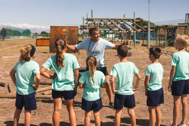 Eine Gruppe kaukasischer Jungen und Mädchen hört an einem sonnigen Tag in einem Bootcamp den Anweisungen eines kaukasischen Fitnesstrainers zu, steht und schenkt ihm Aufmerksamkeit, während er gestikuliert und erklärt. — Stockfoto