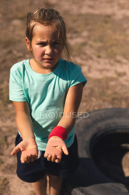 Портрет Кавказької дівчини в чоботному таборі в сонячний день, тримаючи і показуючи їй поранені руки з наклеєними штукатурками на долонях, одягнені в червону стрічку, зелену футболку і чорні шорти — стокове фото