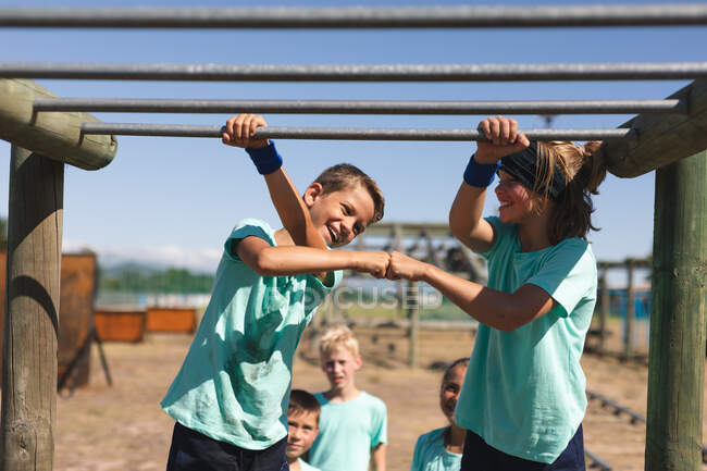 Two smiling Caucasian boys at a boot camp on a sunny day, standing beside each other holding on to the monkey bars, smiling and fist bumping, wearing green t shirts and black shorts — Stock Photo