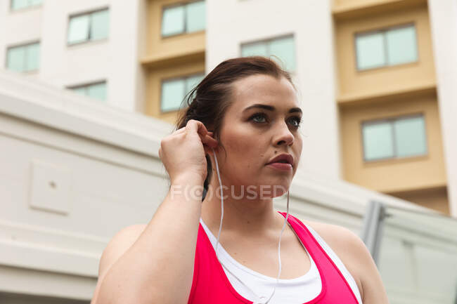 Kurvige kaukasische Frau mit langen dunklen Haaren, die in Sportkleidung in einer Stadt turnt, ihre Kopfhörer aufsetzt und im Hintergrund moderne Gebäude sieht — Stockfoto