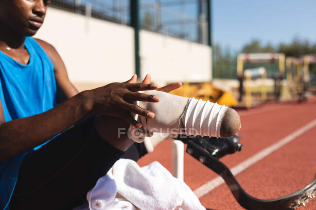 Mid section of fit, mixed race disabled male athlete at an outdoor sports stadium, sitting on race track preparing running blades before workout. — Stock Photo