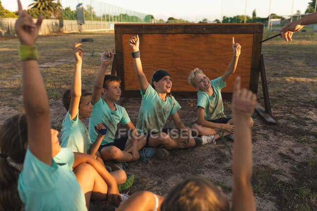 Un groupe de garçons et de filles caucasiens dans un camp d'entraînement par une journée ensoleillée, assis sur l'herbe, souriant et levant la main, se portant volontaires pour un entraîneur de conditionnement physique invisible — Photo de stock