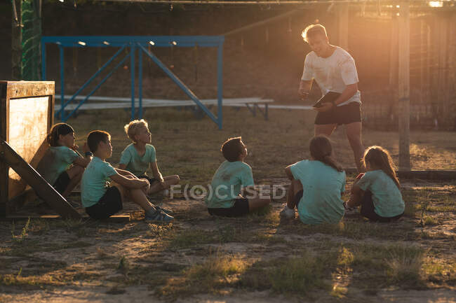 Група кавказьких хлопчиків і дівчаток слухають інструкції від кавказького тренера чоловічої фітнес-команди в таборі чобіт в сонячний день, сидячи на траві, звертаючи увагу на нього, поки він стоїть говорить — стокове фото
