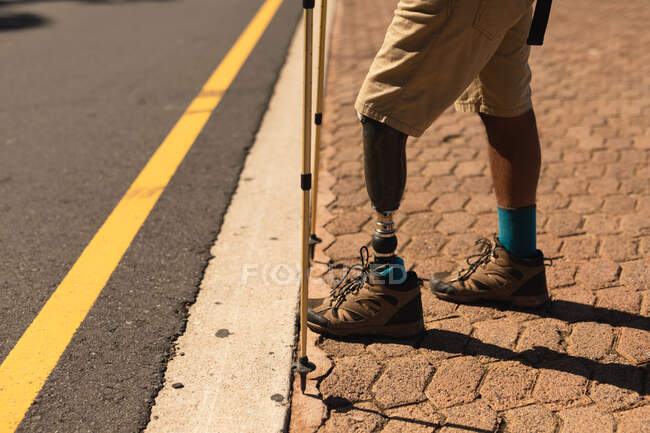 Low section of a fit, disabled male athlete with prosthetic leg, enjoying his time on a trip to the mountains, hiking with sticks, standing on the road. Active lifestyle with disability. — Stock Photo