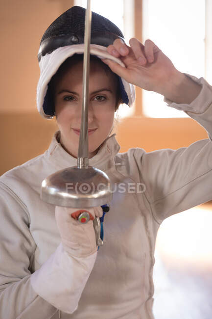 Portrait of Caucasian sportswoman wearing protective fencing outfit during a fencing training session, looking at camera and smiling, holding an epee. Fencers training at a gym. — Stock Photo