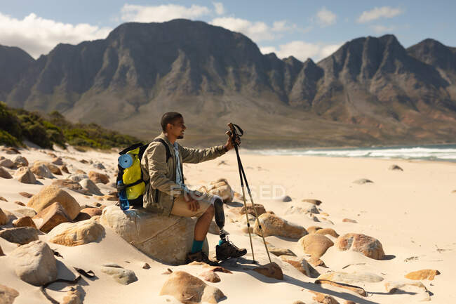 A fit, disabled mixed race male athlete with prosthetic leg, enjoying his time on a trip to the mountains, hiking with sticks, resting on the beach. Active lifestyle with disability. — Stock Photo