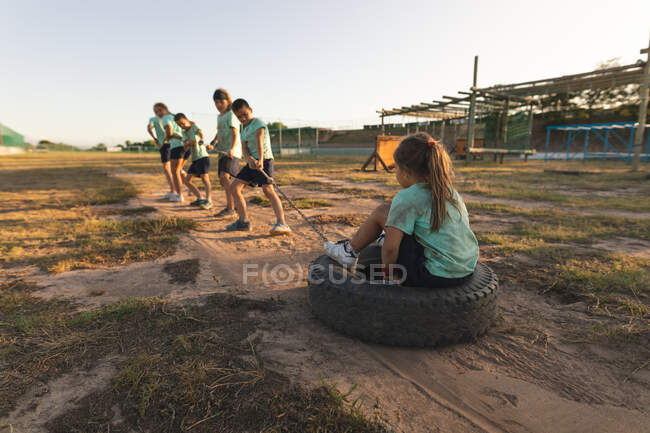 Un grupo de niños y niñas caucásicos con camisetas verdes y pantalones cortos negros en un campamento de entrenamiento en un día soleado, tirando de una chica sentada en un neumático junto con una cuerda - foto de stock