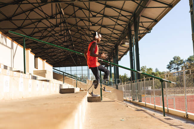 In forma, atleta maschio disabile di razza mista in uno stadio sportivo all'aperto, che corre giù per le scale sugli spalti indossando cuffie e lame da corsa. Disabilità atletica allenamento sportivo. — Foto stock