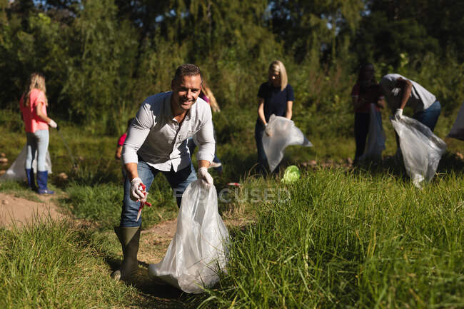 Портрет кавказского волонтера по охране природы, очищающего реку в сельской местности, его друзья собирают мусор на заднем плане. Экология и социальная ответственность в сельской местности. — стоковое фото