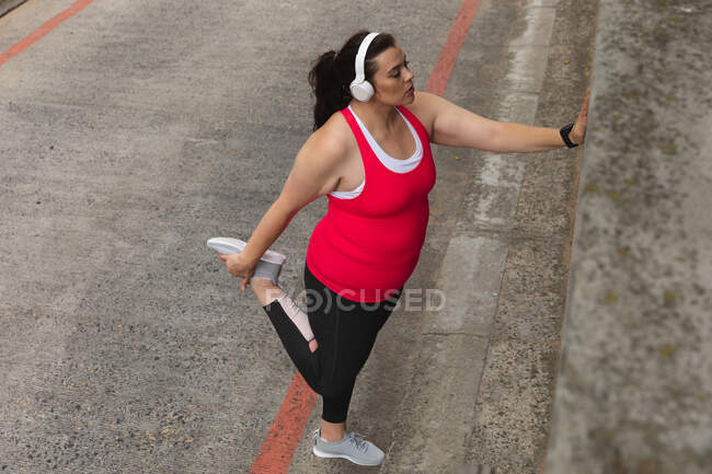 Mujer curvilínea caucásica con el pelo largo y oscuro usando ropa deportiva haciendo ejercicio en una ciudad con auriculares puestos, sosteniendo su pie detrás de ella y estirando su pierna, apoyada contra una pared en una pasarela - foto de stock