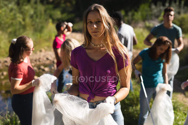 Ritratto di volontaria caucasica che ripulisce il fiume in campagna, i suoi amici che raccolgono spazzatura sullo sfondo. Ecologia e responsabilità sociale nell'ambiente rurale. — Foto stock
