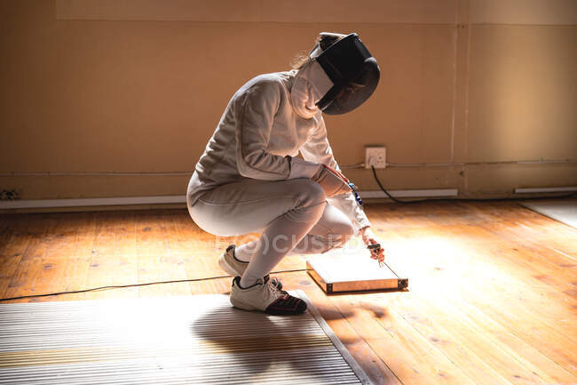 Caucasian sportswoman wearing protective fencing outfit during a fencing training session, preparing the body cord before a duel, squating and checking equipment. Fencers training at a gym. — Stock Photo