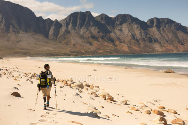 Athlète masculin en forme, handicapé, de race mixte avec une jambe prothétique, profitant de son temps sur un voyage à la montagne, randonnée avec des bâtons, marche sur la plage au bord de la mer. Mode de vie actif avec handicap. — Photo de stock