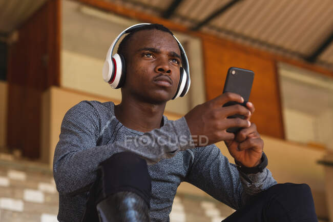 Fit, im Mixed-Rennen behinderter männlicher Athlet in einem Outdoor-Sportstadion, auf der Tribüne sitzend mit Kopfhörern und einem Smartphone mit Laufklingen. Behindertensport. — Stockfoto