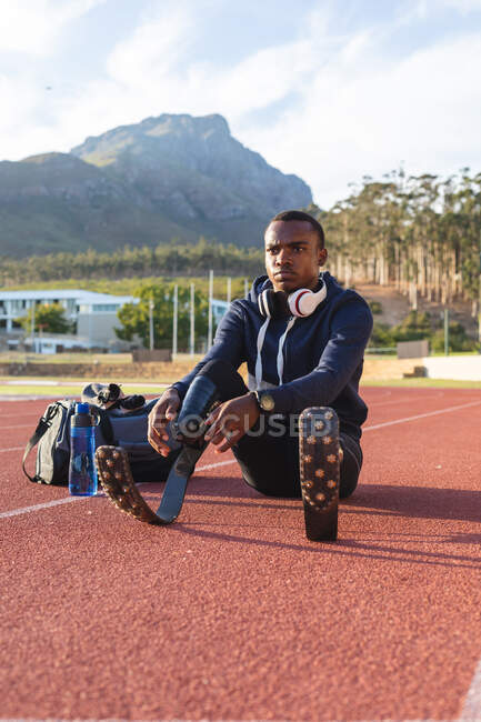 Fit, mixed race disabled male athlete at an outdoor sports stadium, with gym bag and water bottle preparing on race track wearing running blades. Disability athletics sport training. — Stock Photo