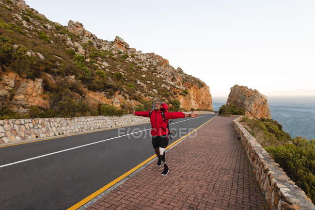Ein fitter, behinderter Mixed-Race-Athlet mit Beinprothese, der seine Zeit bei einem Ausflug in die Berge genießt und mit ausgestreckten Armen auf der Straße am Meer wandert. Aktiver Lebensstil mit Behinderung. — Stockfoto