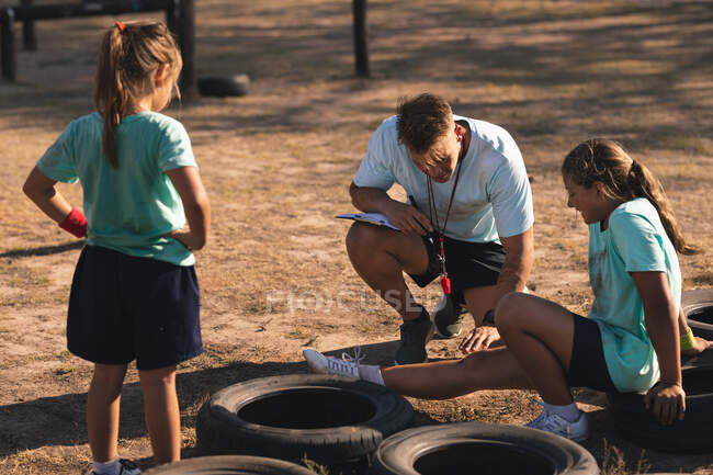 Кавказский тренер по фитнесу, сидящий на корточках и ухаживающий за травмированной ногой белой девушки, сидящей на шине в лагере в солнечный день, с другой девушкой, смотрящей на — стоковое фото