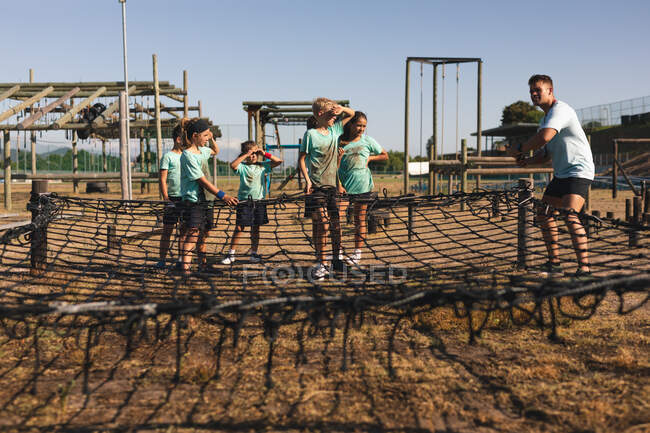 Un allenatore di fitness maschile caucasico istruisce un gruppo di ragazzi e ragazze caucasici che indossano magliette verdi e pantaloncini neri accanto a una rete di corda in un campo di addestramento in una giornata di sole — Foto stock