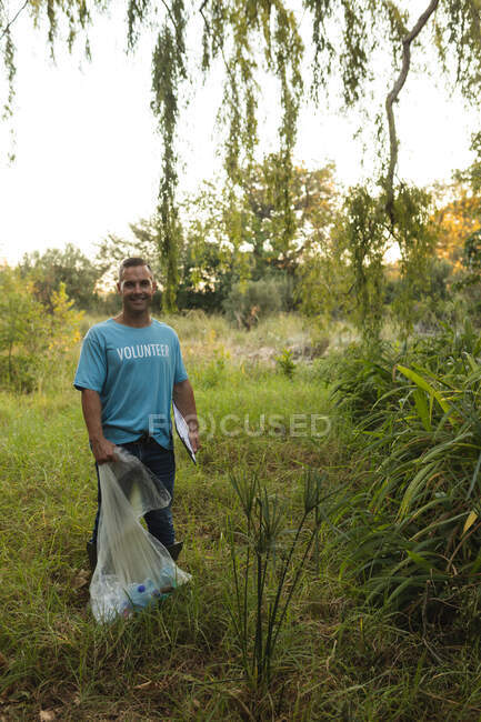 Retrato de feliz hombre caucásico voluntario de conservación limpiando el bosque en el campo, sujetando el portapapeles y la bolsa de basura. Ecología y responsabilidad social en el medio rural. - foto de stock