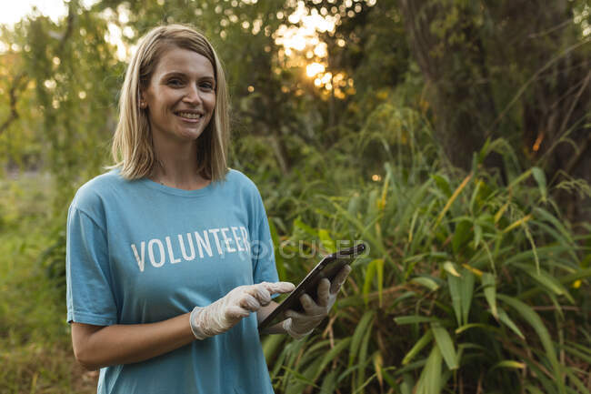 Portrait of happy Caucasian female conservation volunteer cleaning up forest in the countryside, holding digital tablet, smiling to camera. Ecology and social responsibility in rural environment. — Stock Photo